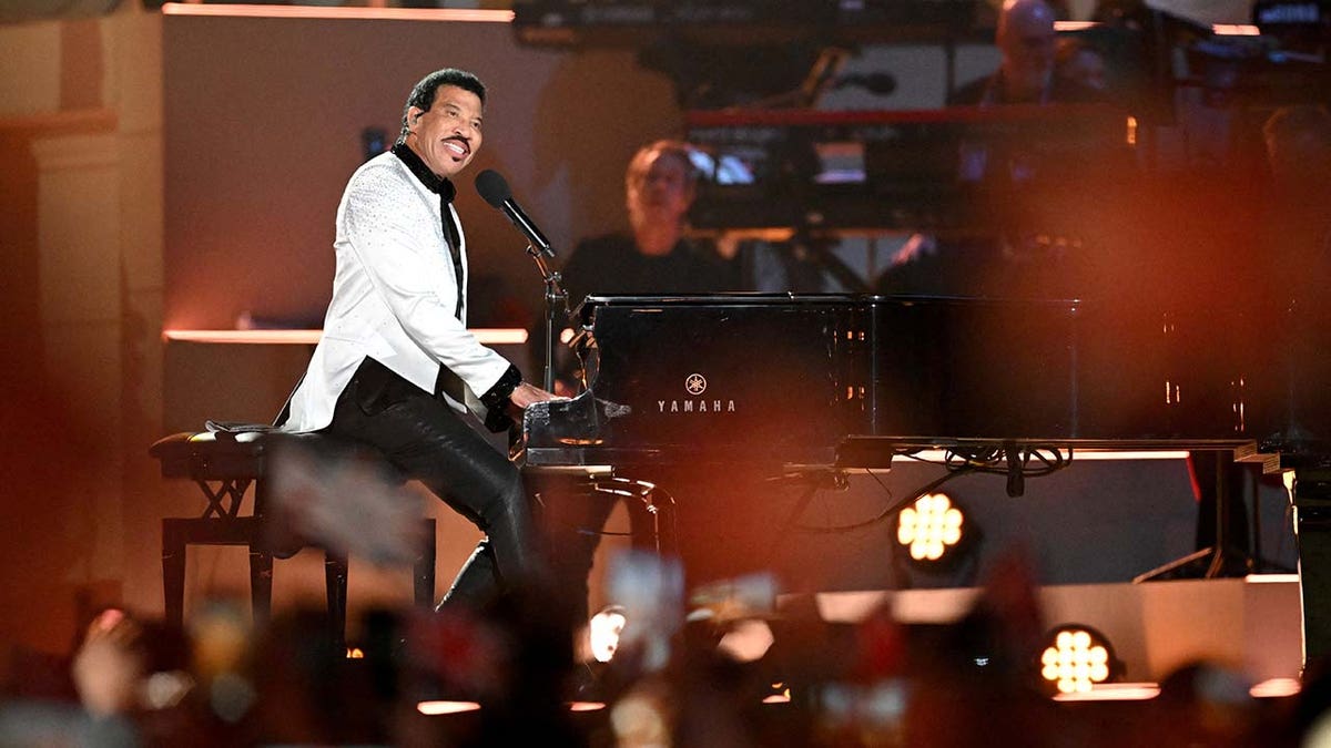 Lionel Richie on stage playing the piano looks out to the crowd at the coronation concert, wearing a white jeweled jacket