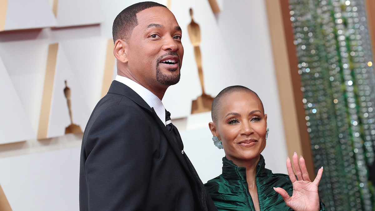 Will Smith looks slightly up on the carpet at the Oscars with Jada Pinkett Smith smiling next to him in a green gown